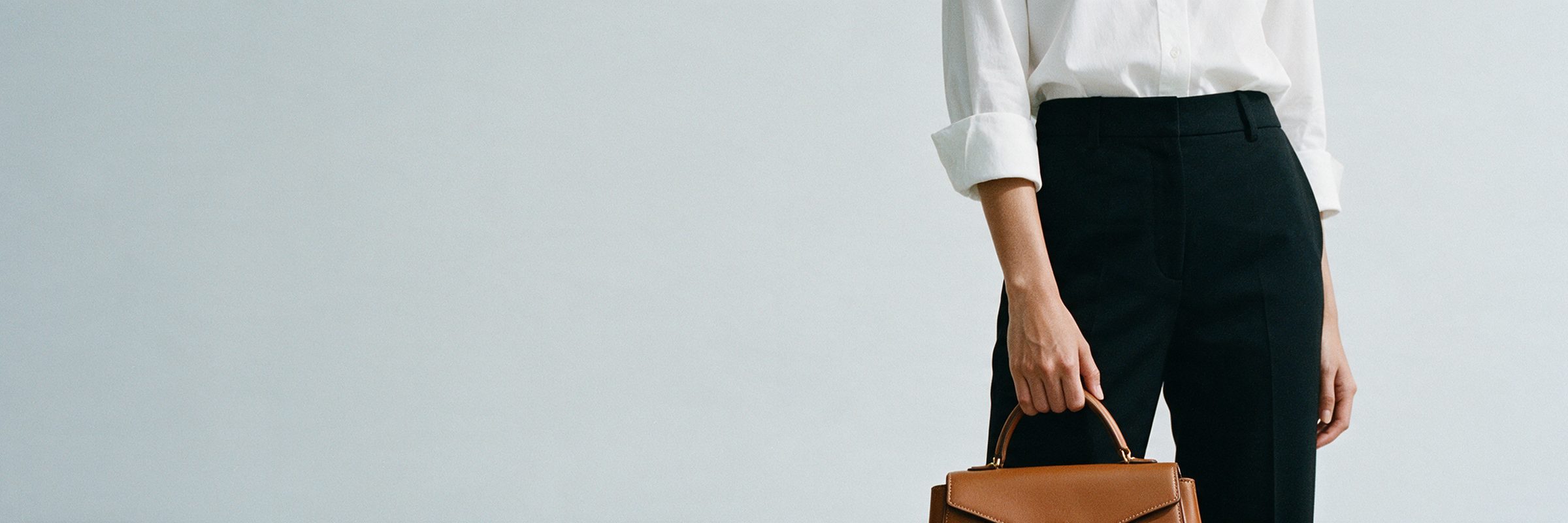 A cropped editorial banner of a figure in a white shirt holding a structured tan handbag