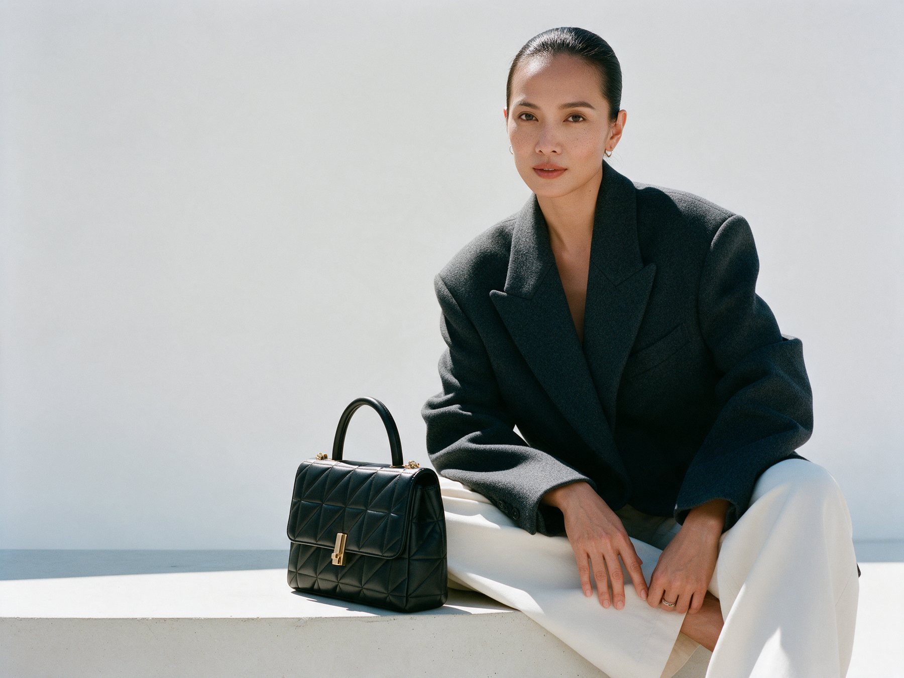 A structured black quilted handbag with gold hardware beside a seated model against a bone-white studio backdrop
